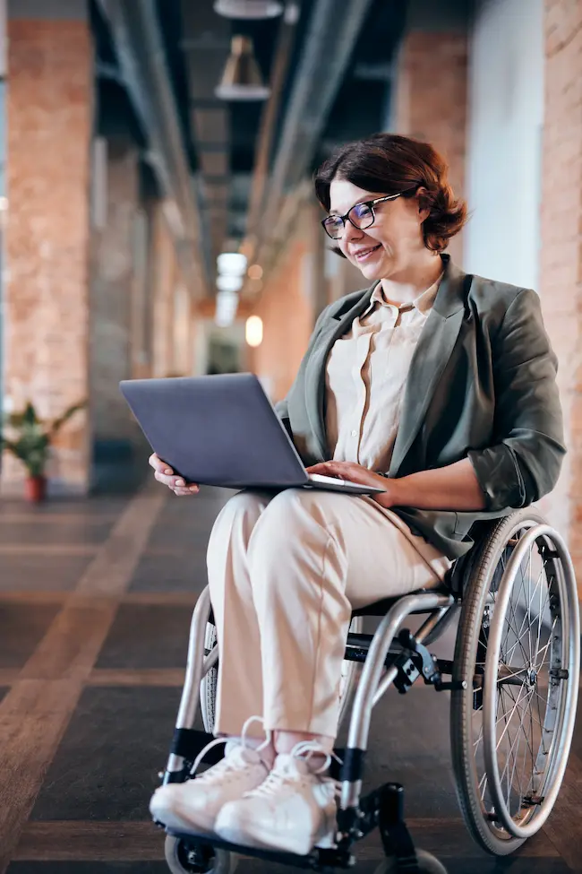 Eine lächelnde Frau im Rollstuhl sitzt in einem modernen Bürogebäude und arbeitet an ihrem Laptop. Sie trägt eine Brille und einen schicken Blazer, was auf eine berufliche Umgebung hindeutet.